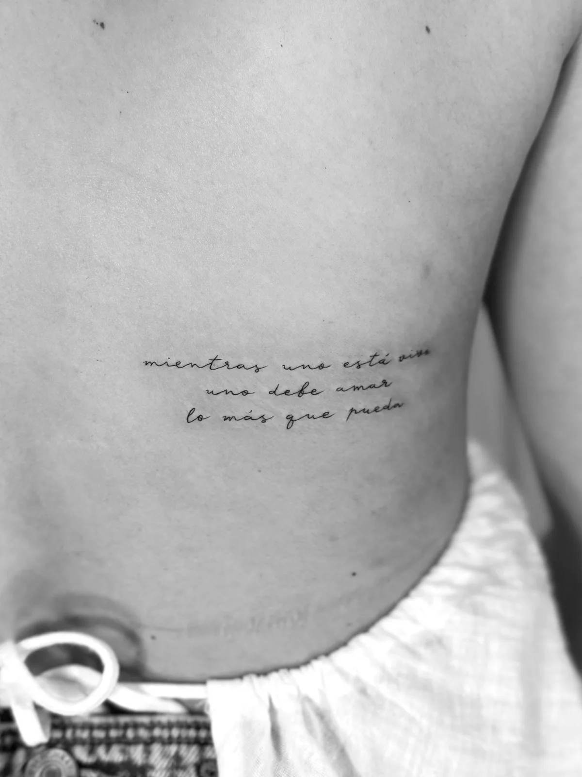 Fine line lettering tattoo 'mientras uno está vivo, uno debe amar lo más que pueda' (Bad Bunny) in 3 cursive lines on the side of the torso — Sinkply Madrid