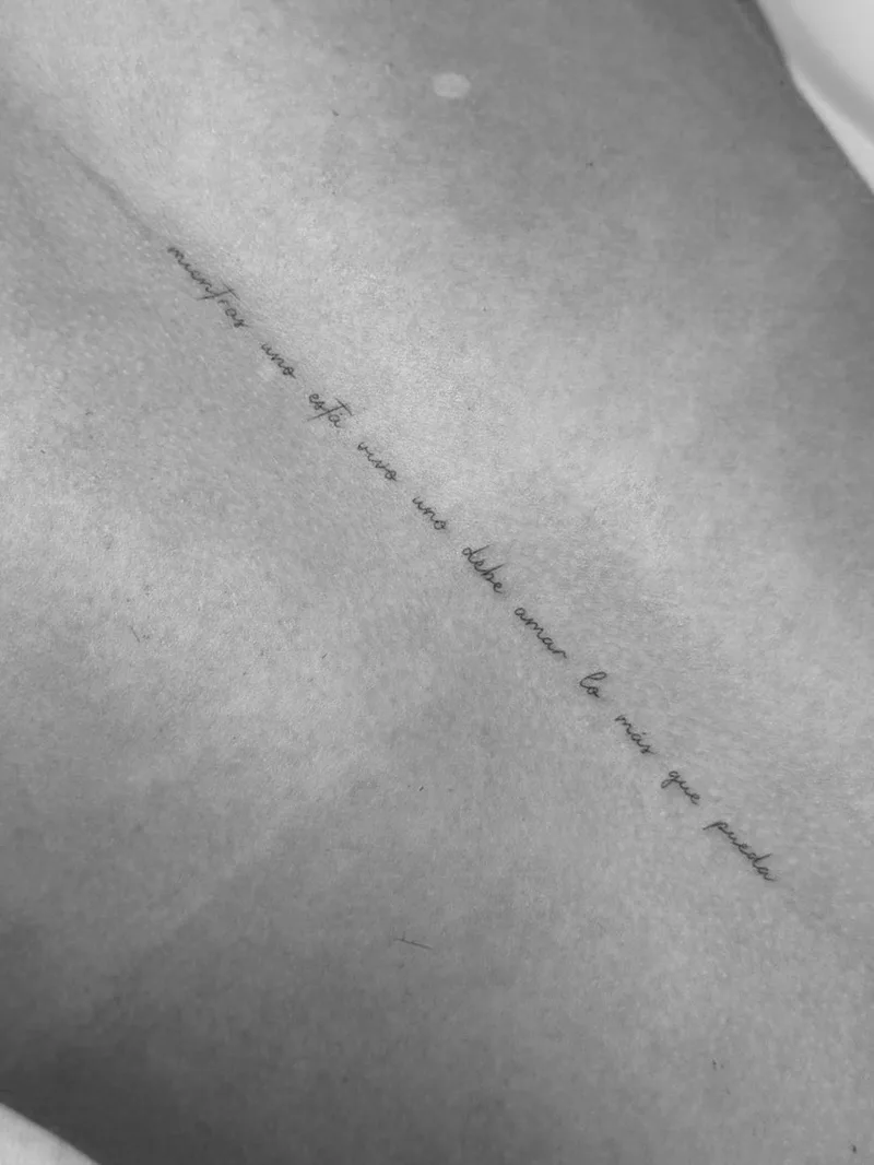 Fine line lettering tattoo of the Bad Bunny lyric “mientras uno está vivo uno debe amar lo más que pueda” in delicate script down the back — Sinkply Madrid