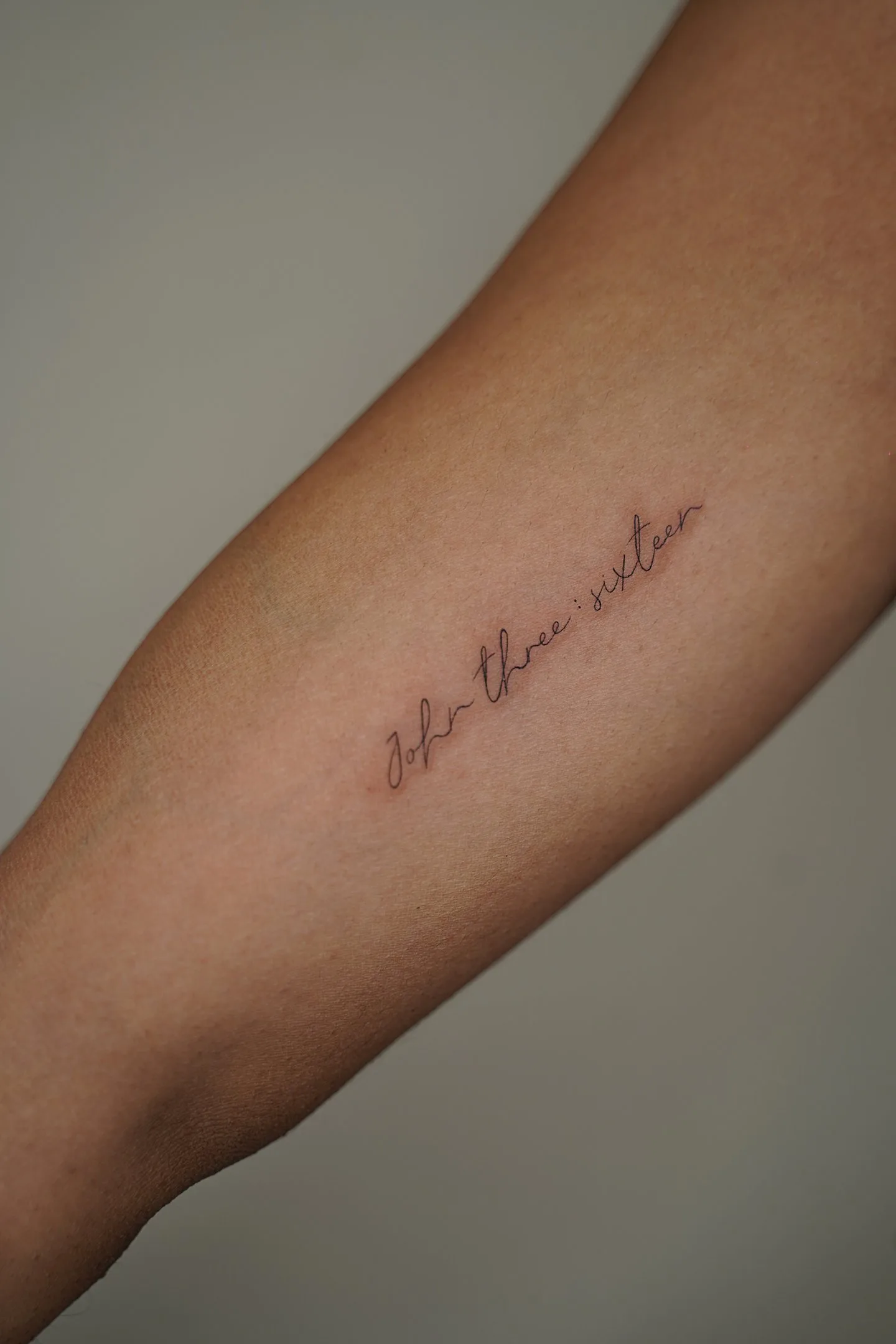 Tatuaje fineline en Madrid de frase John three sixteen en cursiva fina diagonal en el antebrazo