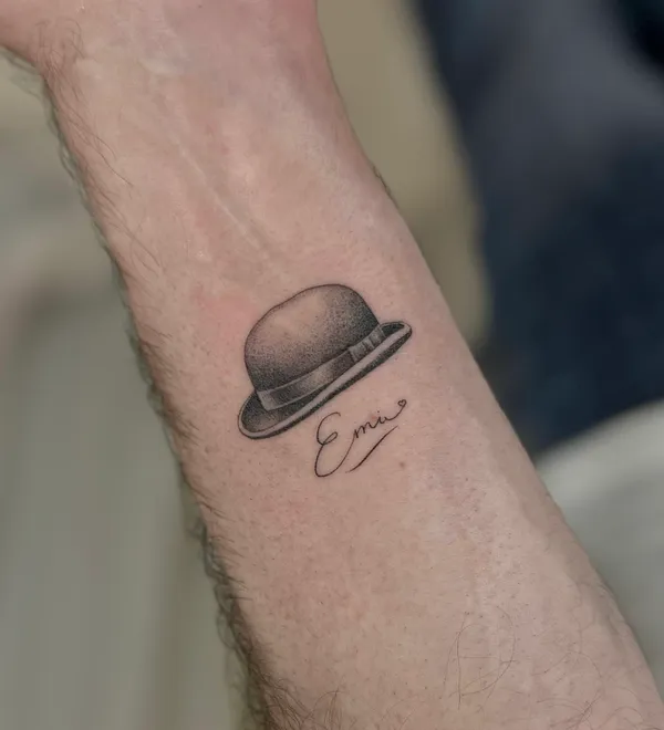 Tatuaje de un sombrero bombín en microrealismo con un nombre en el antebrazo, por Oriana en Sinkply Madrid